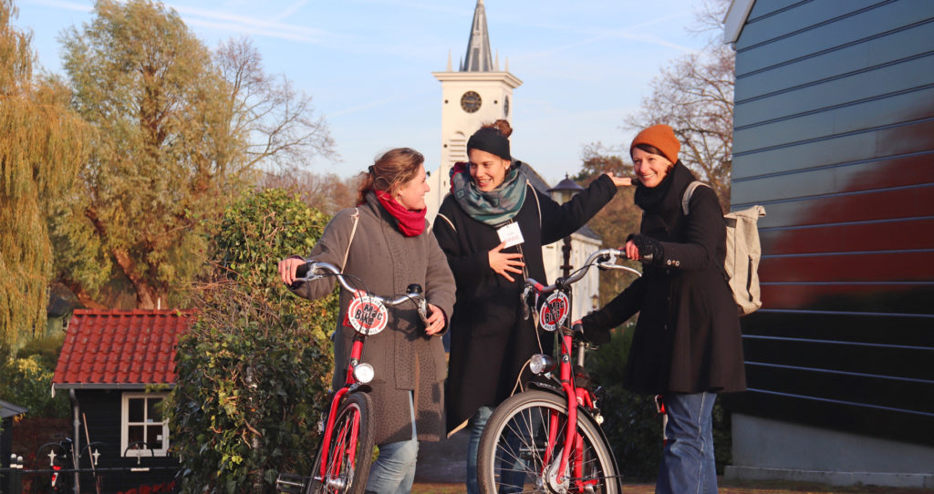 Amsterdamliebe Bike Tour Noord Tour participants stand with tour guide Hanna next to a little house with the Schellingwouderkerk in the background